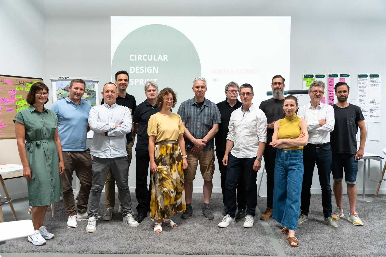 Dreizehn Personen stehen als Gruppe zusammen und lächeln in die Kamera, im Hintergrund ist die Projektion eines Beamers und verschiedene Workshop-Boards/Pinnwände zusehen